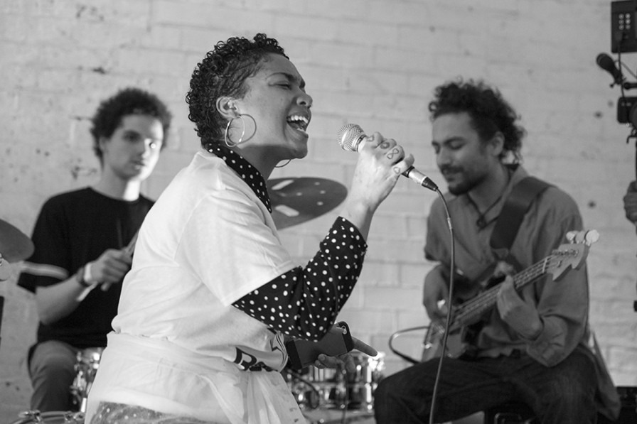 Photo from Brighter Sound a young woman passionately sings into a microphone. her hair is short and curly and there are two band members seen in the background.